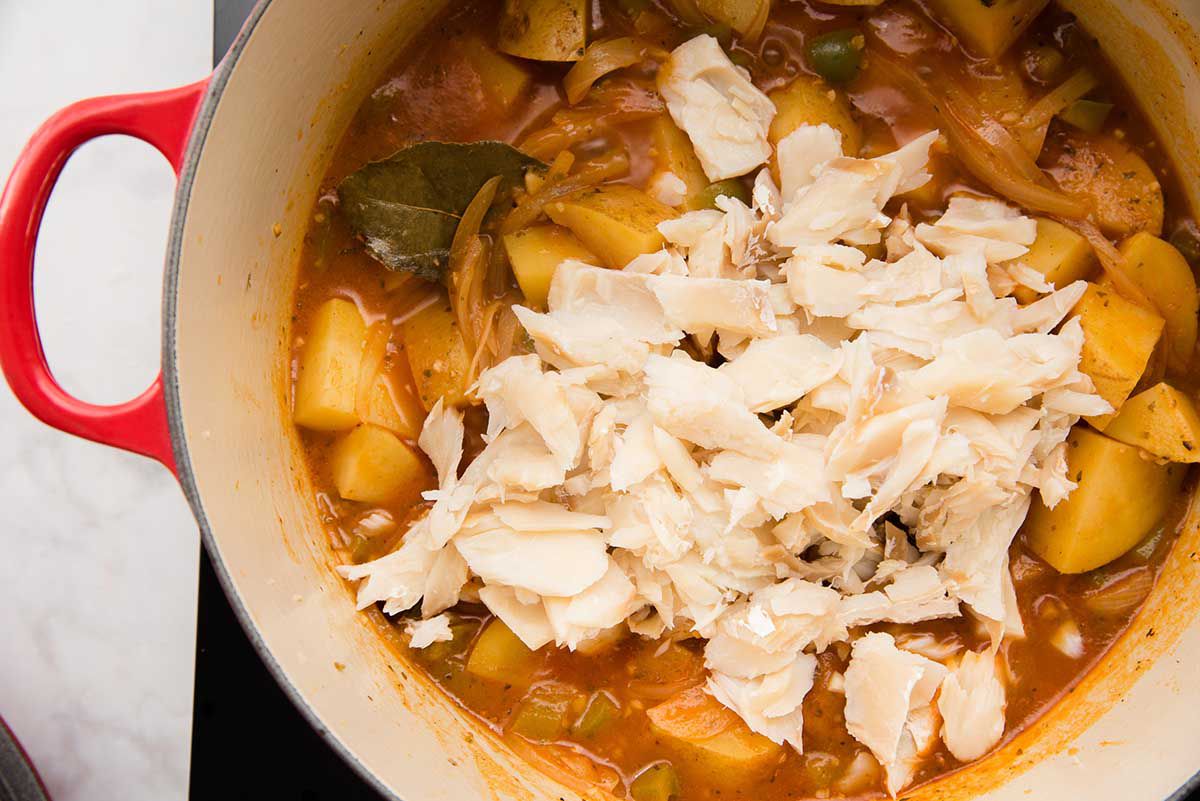 A dutch over with flaked cod on top of a stew of potatoes, bay leaf and a red broth.