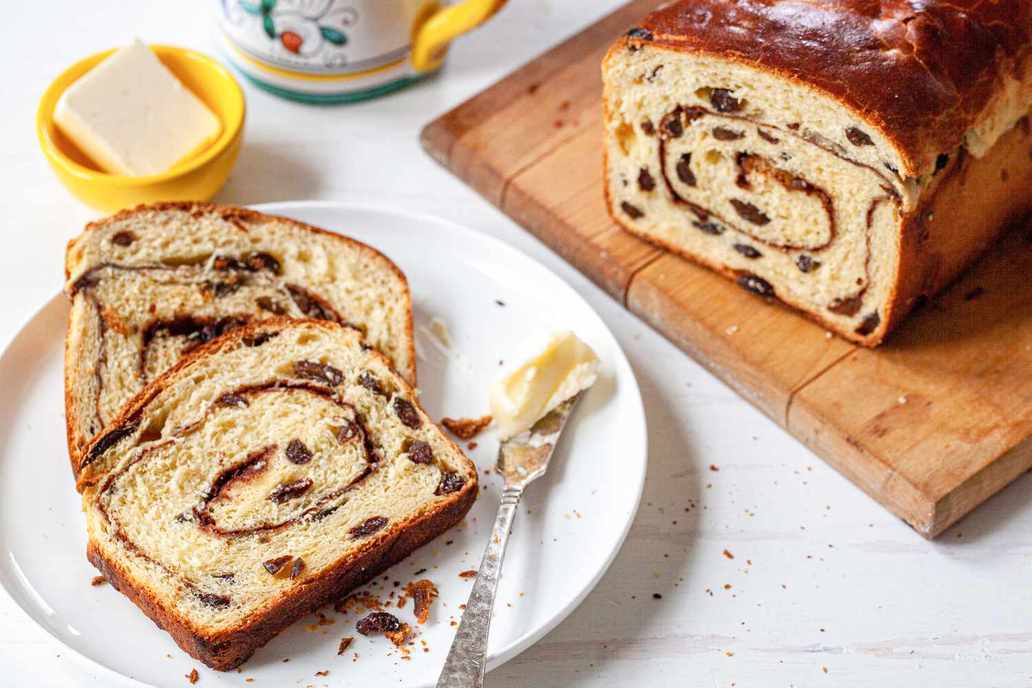 Slices of Cinnamon Swirl Raisin Brioche Loaf on a Small Plate with Some Butter on a Butter Knife, Next to a Cutting Board with the Remaining Loaf and a Small Bowl with More Butter