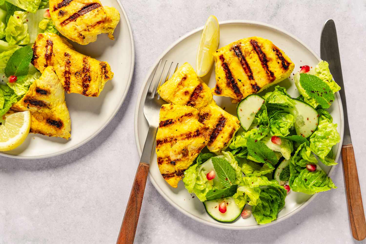 Grilled chicken breasts and fresh salad on plates, garnished with lemon slices and pomegranate seeds