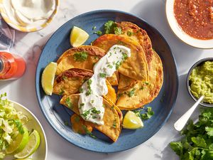 Bowl of cheesy baked tacos on a plate topped with sour cream and cilantro at a table setting with a bowl of lime wedges and shredded lettuce, bowl of sour cream, bowl of salsa, bowl of salsa, and a bunch of cilantro