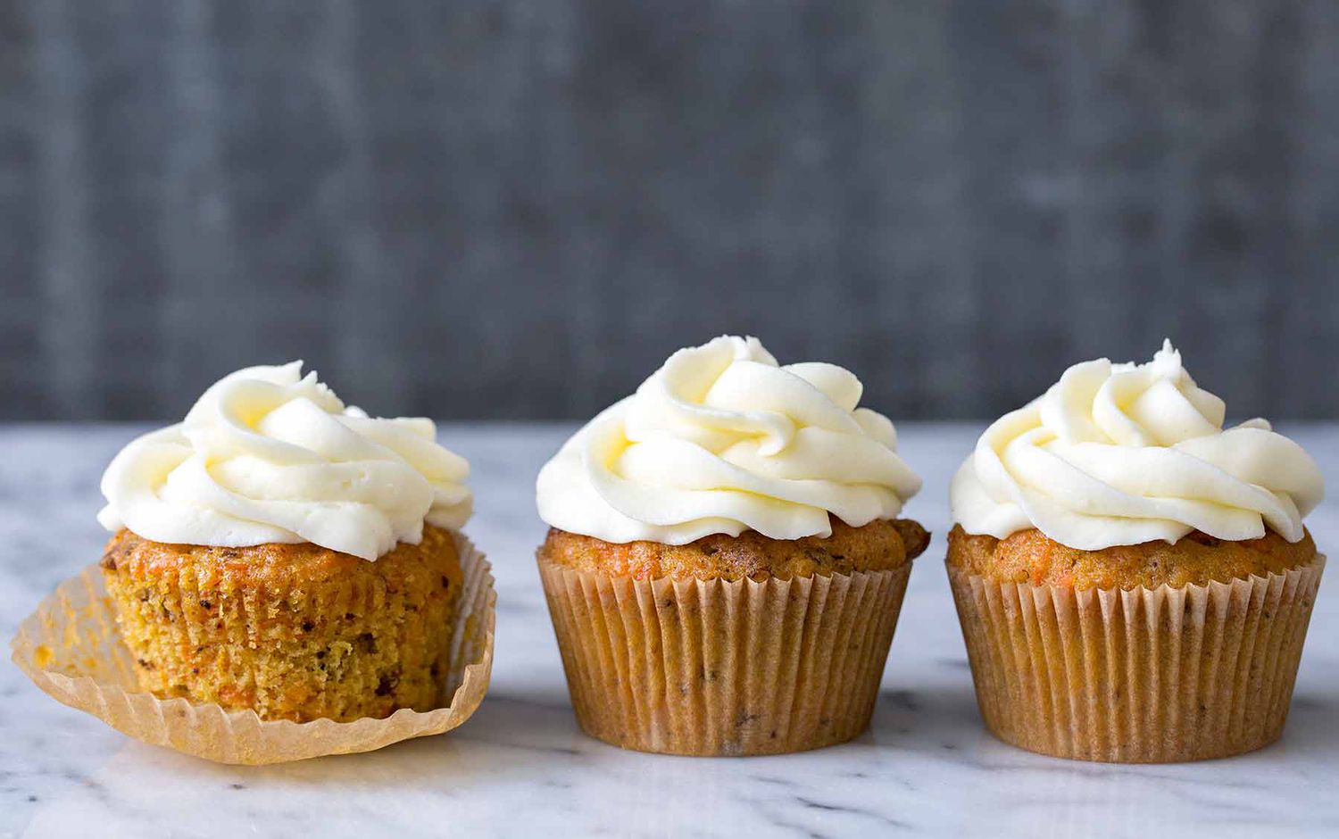 Three Carrot Cake Cupcakes topped with cream cheese frosting, one of them is partially unwrapped