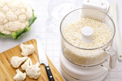 lidless food processor with processed cauliflower in it, head of cauliflower on the side and a few cauliflower florets on a cutting board with a knife