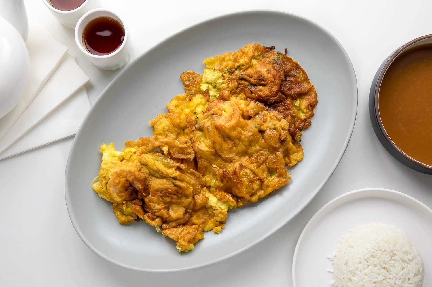Egg Foo Young on a Plate Next to a Plate with Rice, a Pot of Sauce, and Small Cups with Tea 