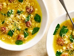 Two bowls of soup with pasta spinach meatballs and grated cheese topped with ground pepper