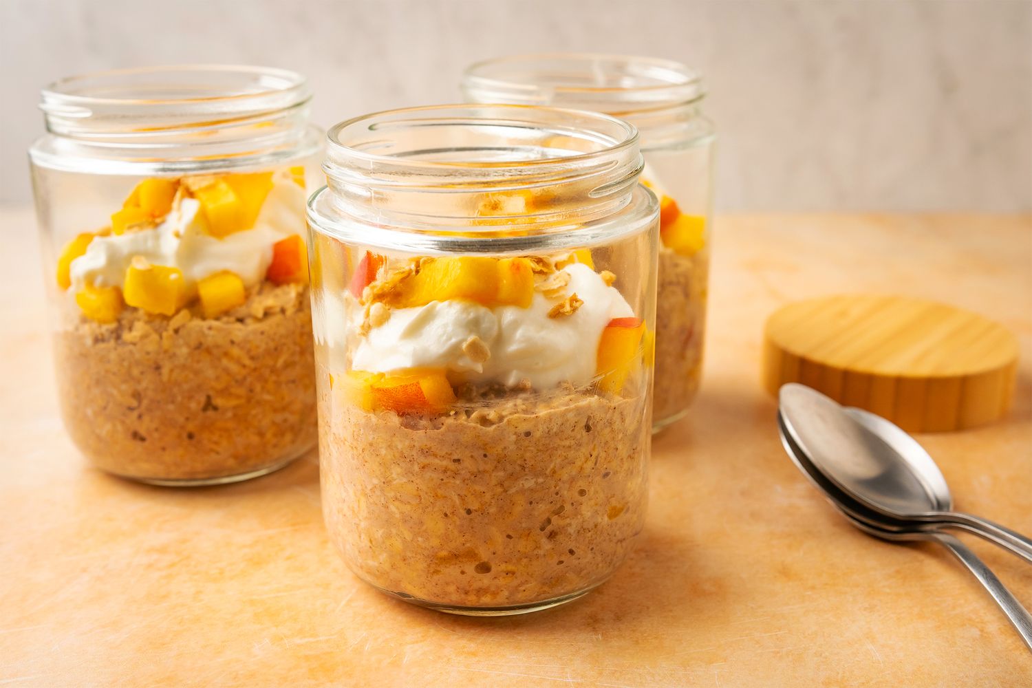 Three jars of peach cobbler overnight oats topped with whipped cream and peach chunks set on a countertop with spoons nearby