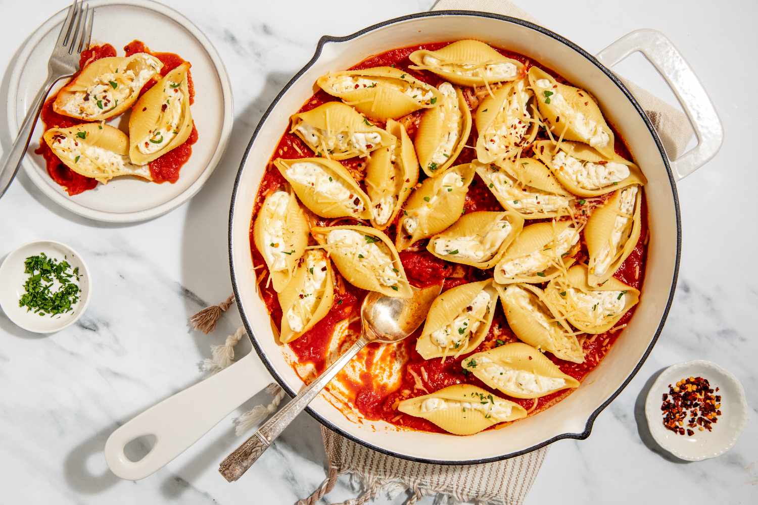Stovetop Stuffed Shells in a large pan with a large serving spoon - small dish with crushed red pepper and small dish with chopped parsley, one small plate with one serving and a fork