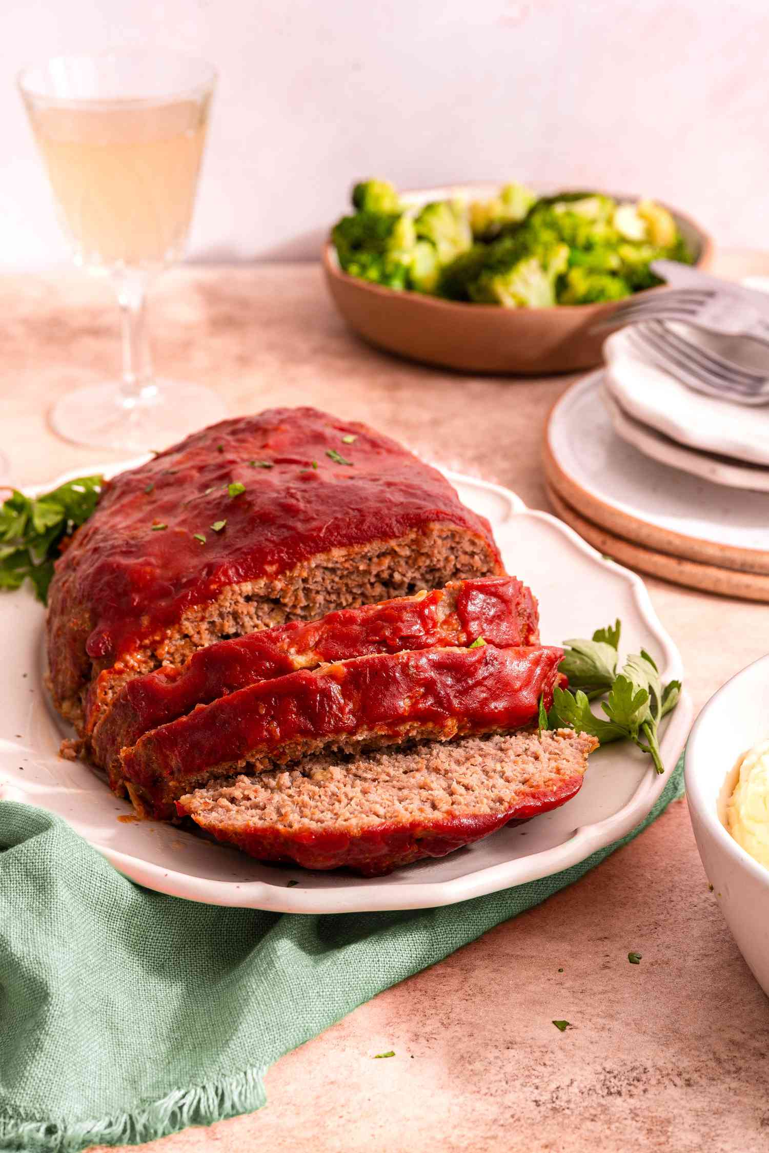 Easy Meatloaf With Some of It Cut Into Slices on a Platter and Sitting on a Sage Table Napkin, and in the Surroundings, a Stack of Plates With Cutlery, a Bowl of Mashed Potatoes, a Bowl of Broccoli, and a Glass of Wine