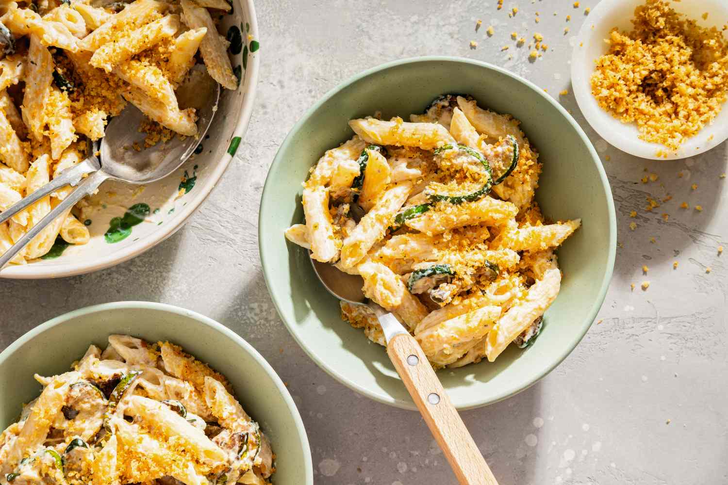 Bowls of pasta with ricotta and zucchini served on a table with a small bowl of breadcrumbs