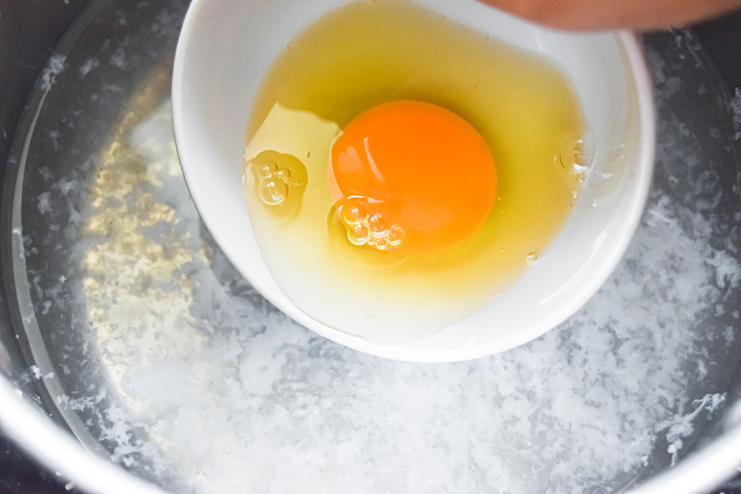 White bowl with a cracked egg inside being added to simmering water 