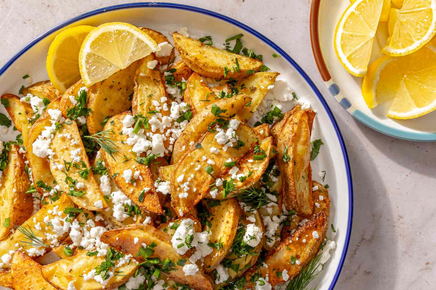 Overhead shot of a serving plate with Greek-style baked potato wedges, garnished with crumbled feta, dill and lemon wedges