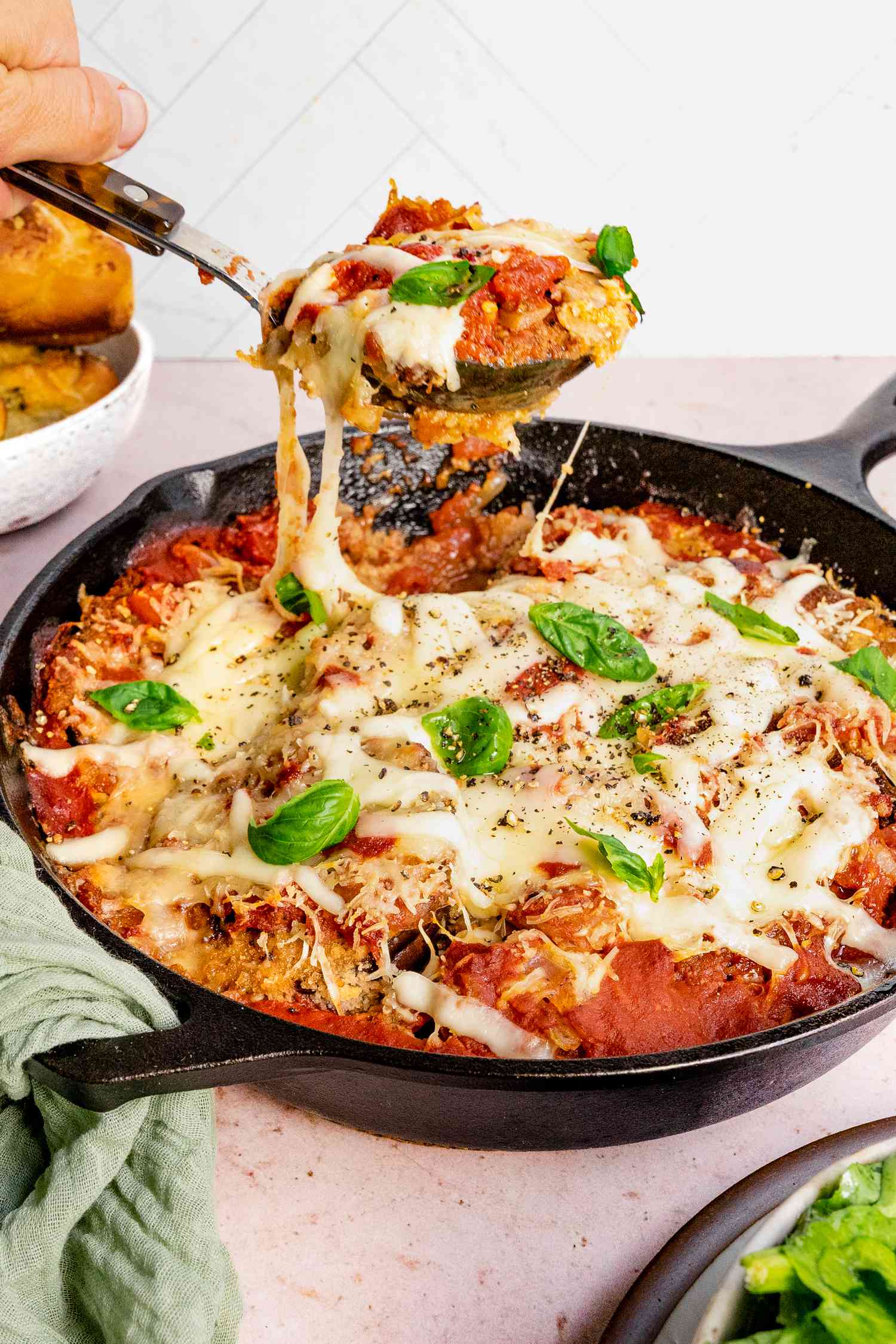 Spoon Lifting a Portion of Skillet Eggplant Topped With Fresh Basil from a Cast Iron Skillet, and in the Surroundings, a Mint Kitchen Linen on the Handle of the Cast Iron Skillet and a Bowl of Salad Greens 