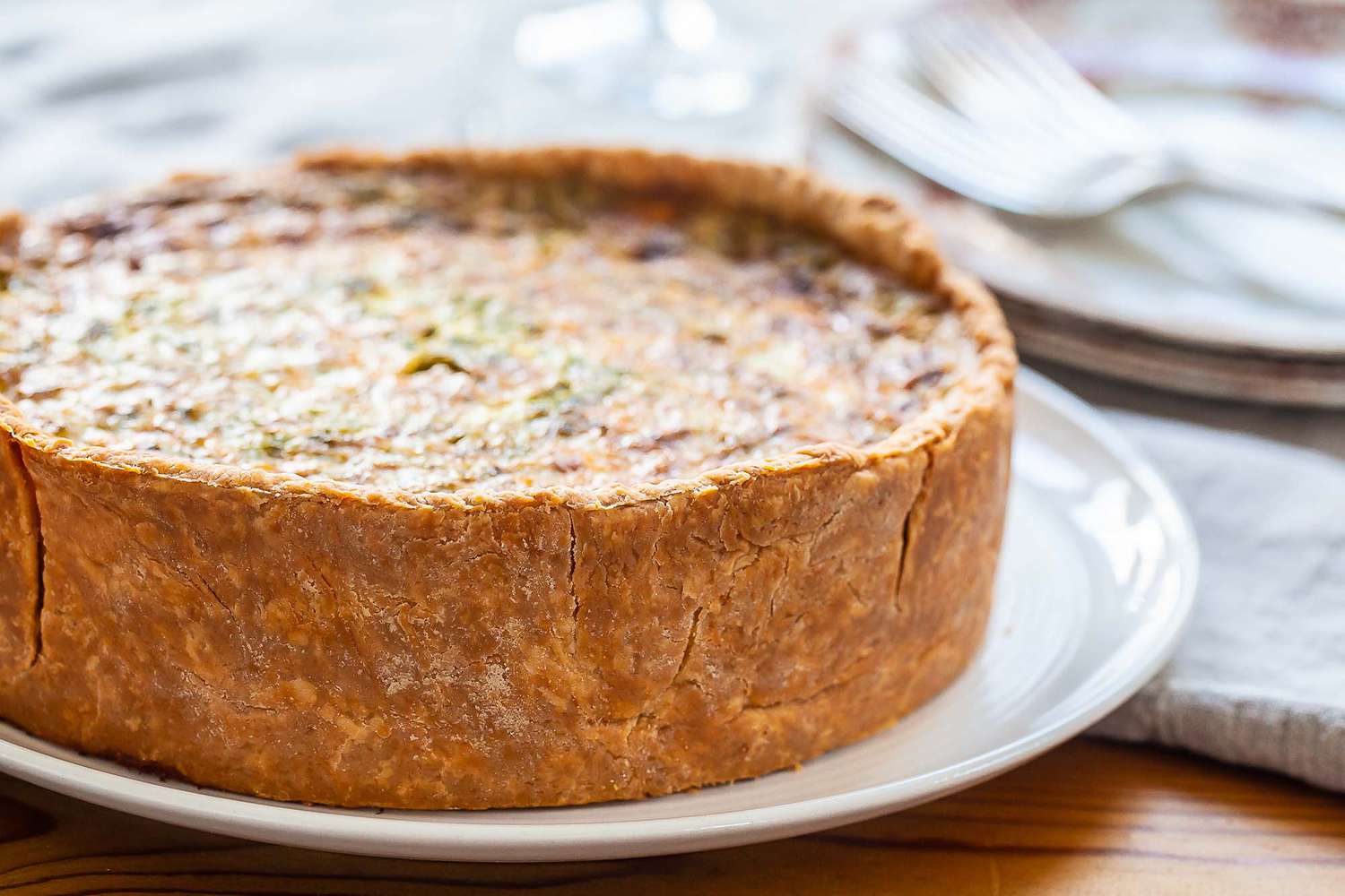 A mile high quiche is on a white platter. The crust is golden brown as is the filling. Partial view of plates and forks to the upper left of the quiche.