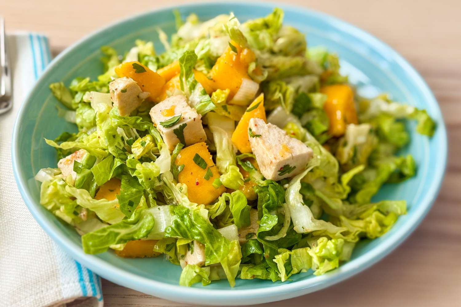 A close-up of a salad on a plate featuring lettuce, mango chunks, herbs, and chicken slices, placed on a table next to a cloth napkin and fork