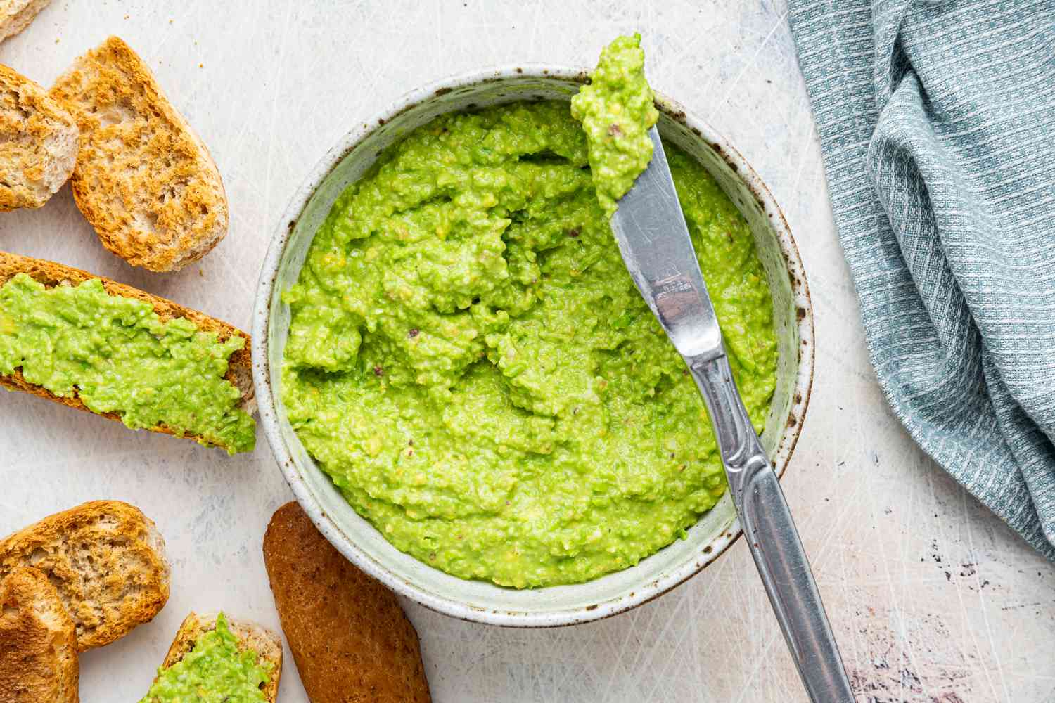 Table Knife With a Bit of Pea Pesto Rested on Top of a Bowl of Pea Pesto Surrounded by Slices of Toasted Bread on the Counter (Some Pieces of Bread Is Smeared With Pea Pesto) and a Blue and White Stripped Kitchen Towel 