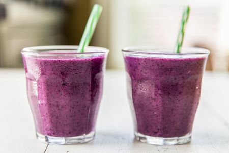 Two glasses of purple smoothies with green straws placed on a white surface