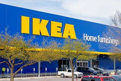 Front view of an IKEA store entrance with a large blue facade and logo cars and trees visible in the foreground