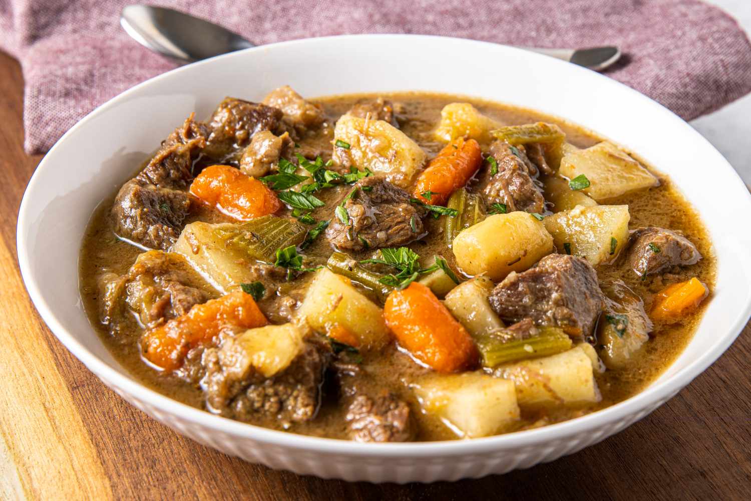 Bowl of beef stew with chunks of beef carrots potatoes celery garnished with herbs