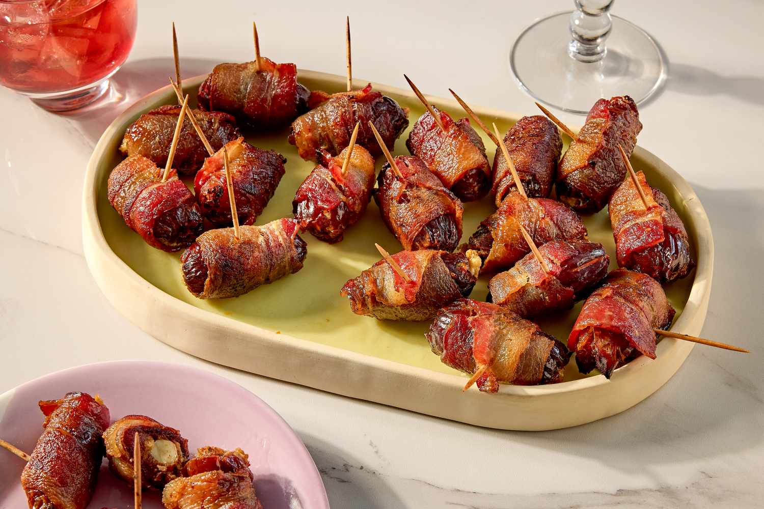 A tray of baconwrapped dates served with toothpicks accompanied by a glass of red drink in the background