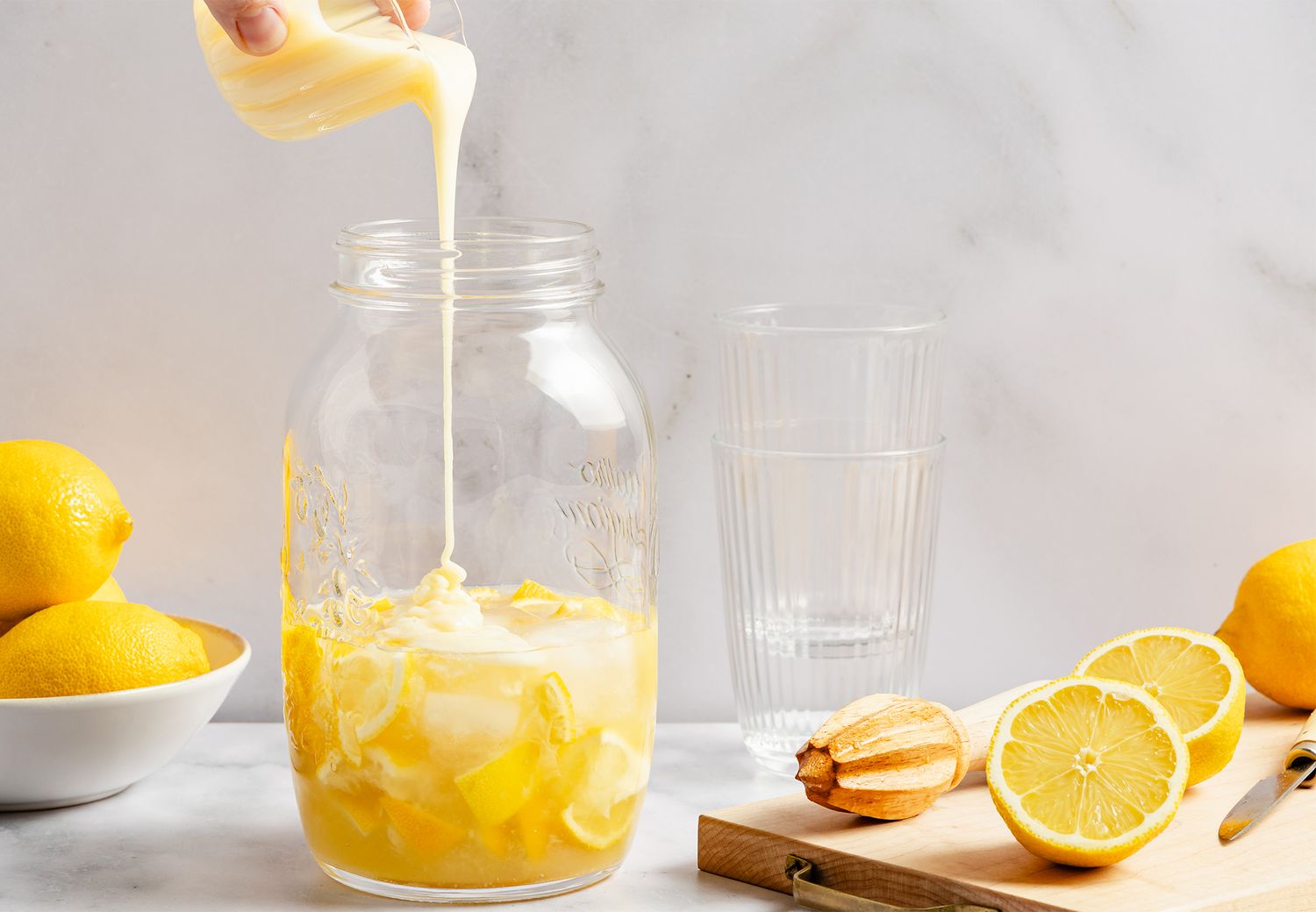 Jar with lemon slices and creamy liquid being poured lemons and a juicer nearby