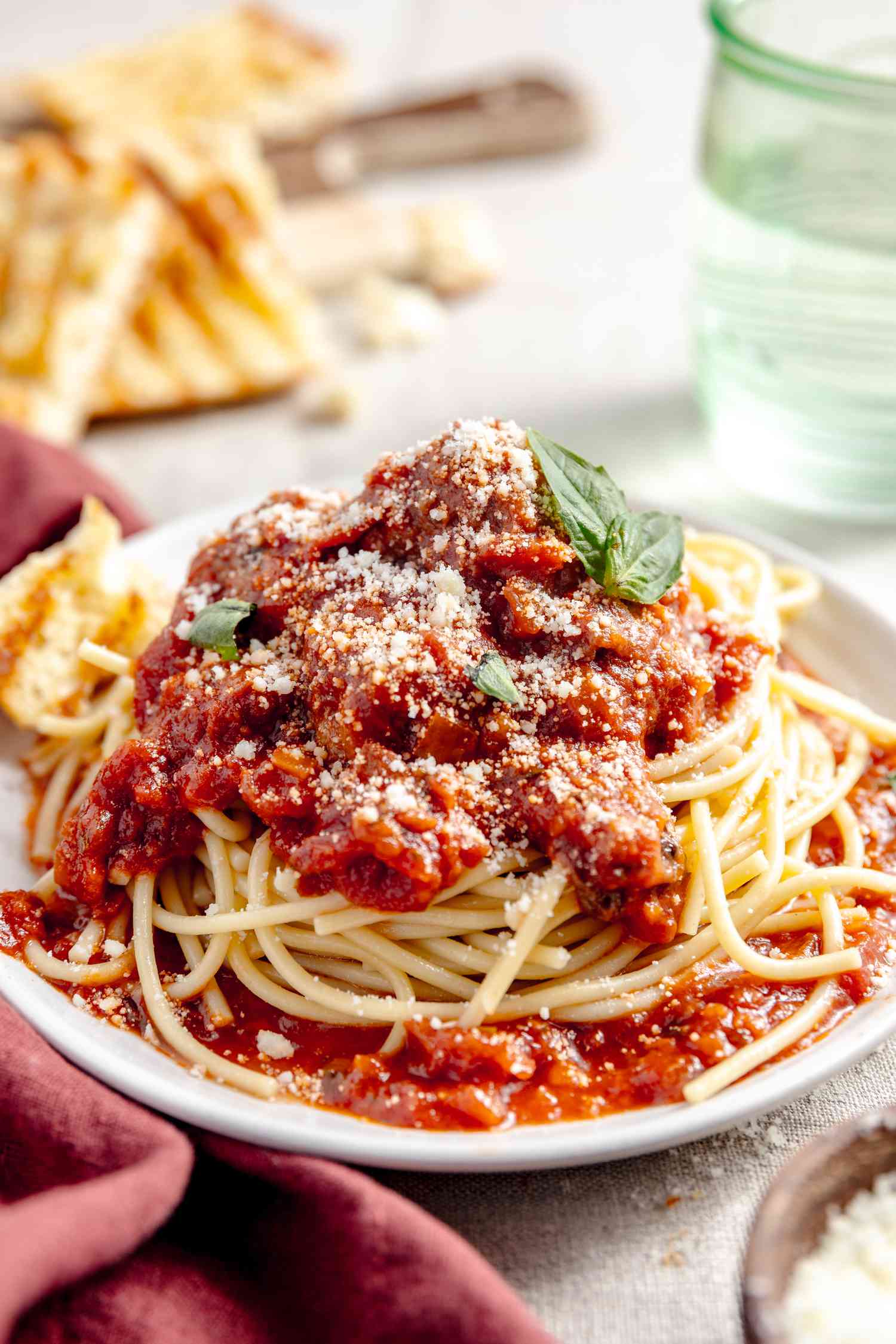 Plate of Spaghetti and Meatballs Topped with Basil, and in the Surroundings, a Glass of Water, Toasted Bread, a Burgundy Table Napkin, and a Bowl of Parmesan