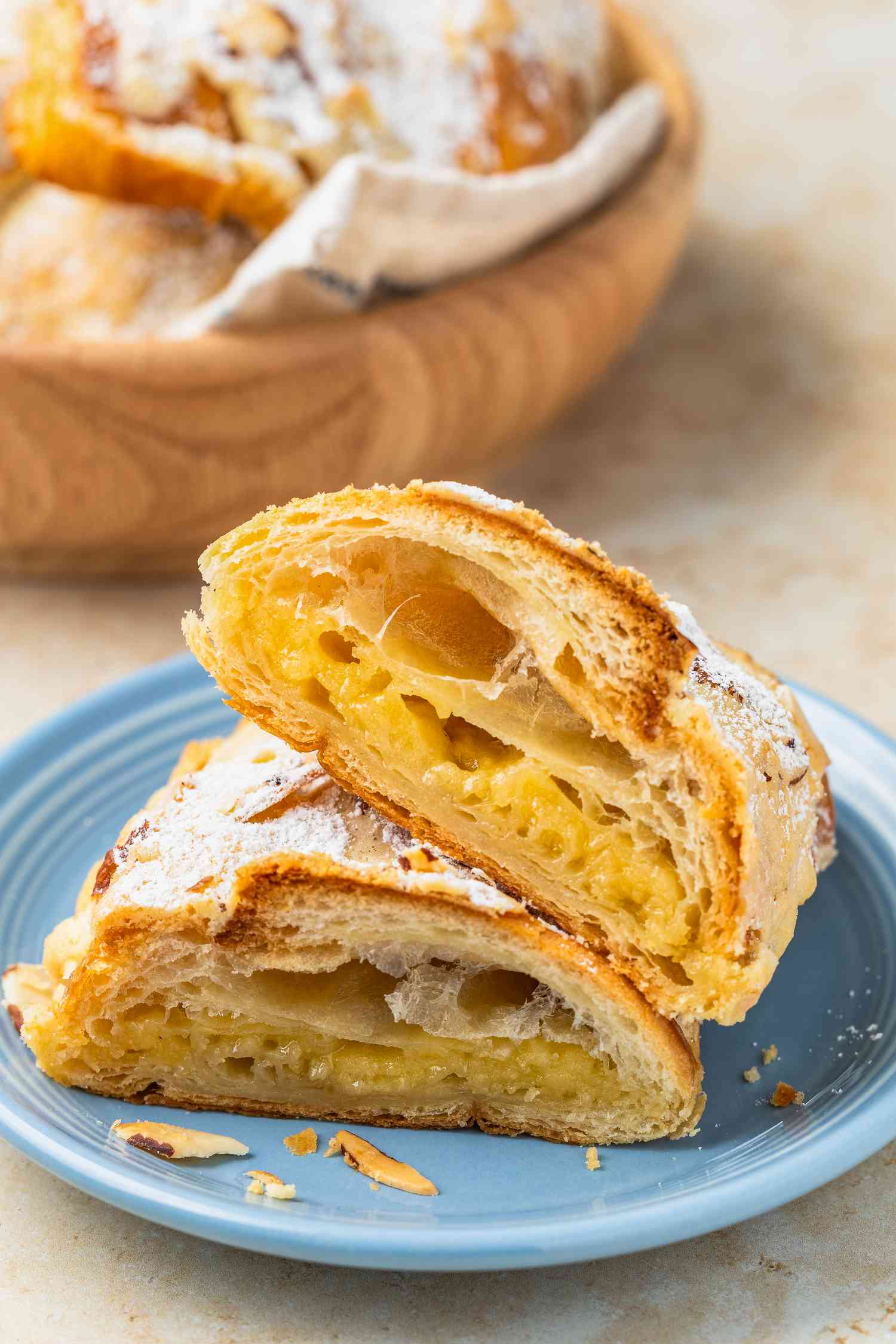 Halved Almond Croissant on a Plate With Cut Side Facing Forward, and Behind It, the More Croissants in Kitchen Towel Covered Wooden Bowl
