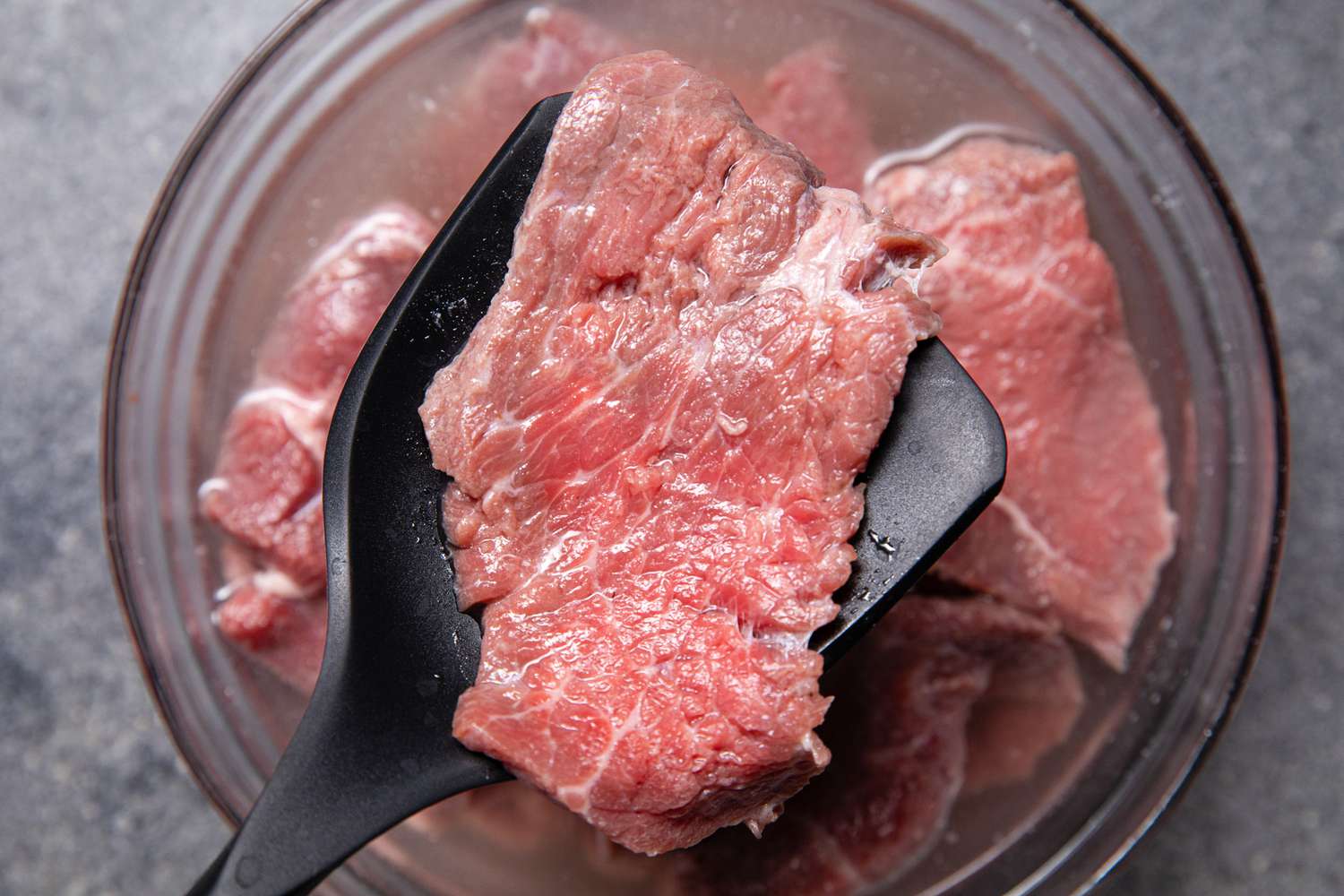 Slice of Beef on a Spatula Raised From a Bowl of Water (the Baking Soda Is Being Rinsed Off Beef Slices)
