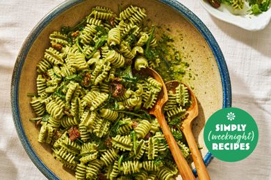 A bowl of rotini pasta with pesto sauce garnished with sundried tomatoes and accompanied by two wooden serving forks