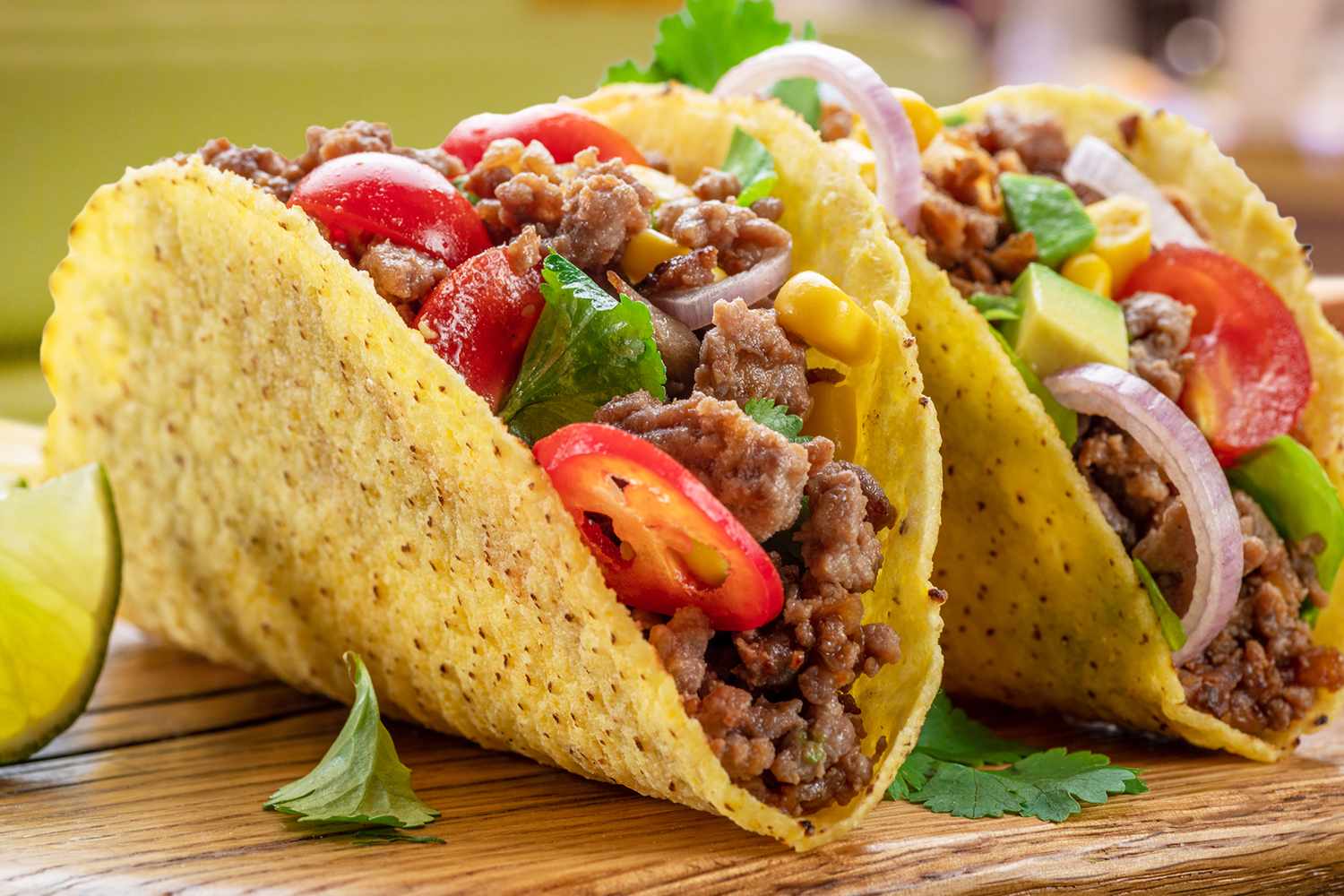 Two tacos on a wooden surface filled with ground beef sliced vegetables and garnishes