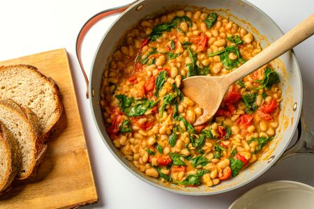 Pan of stewed beans with spinach and tomatoes wooden spoon and sliced bread on a cutting board