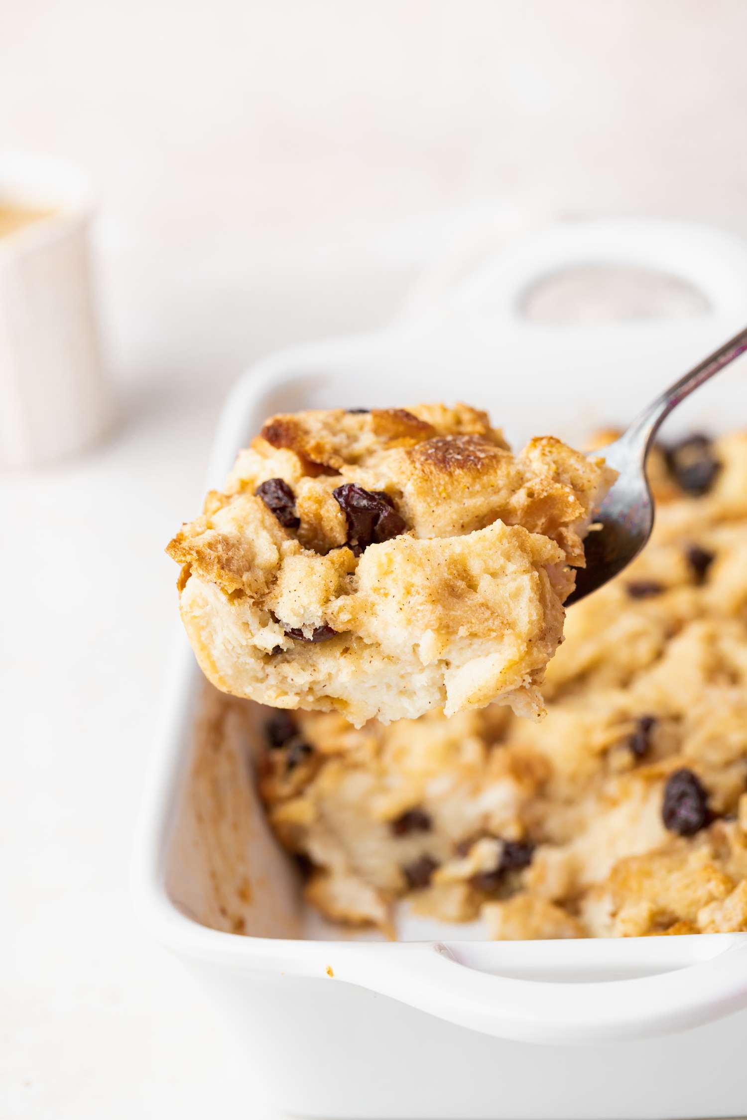 A serving spoon holding a bite of easy bread pudding scooped out of a white casserole dish.