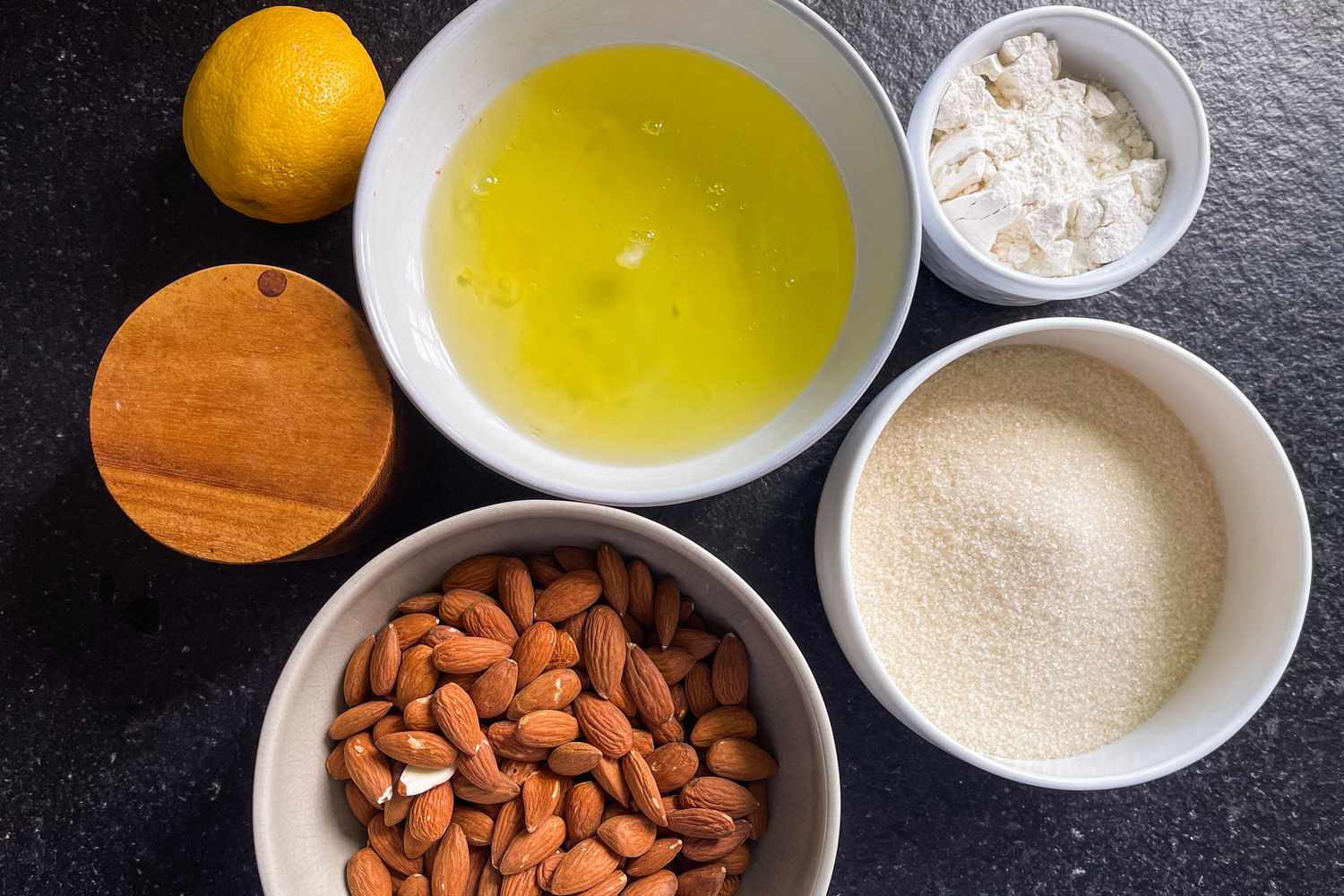 Ingredients for an almond cake arranged on a dark surface, including almonds, sugar, egg whites, flour, and a lemon