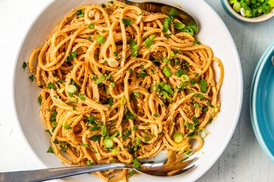 Peanut butter noodles garnished with sesame seeds and scallions in a bowl with serving utensils