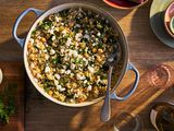 Skillet of chickpea and spinach rice, surrounded by a cutting board of feta and chopped dill, wine glasses, and serving plates