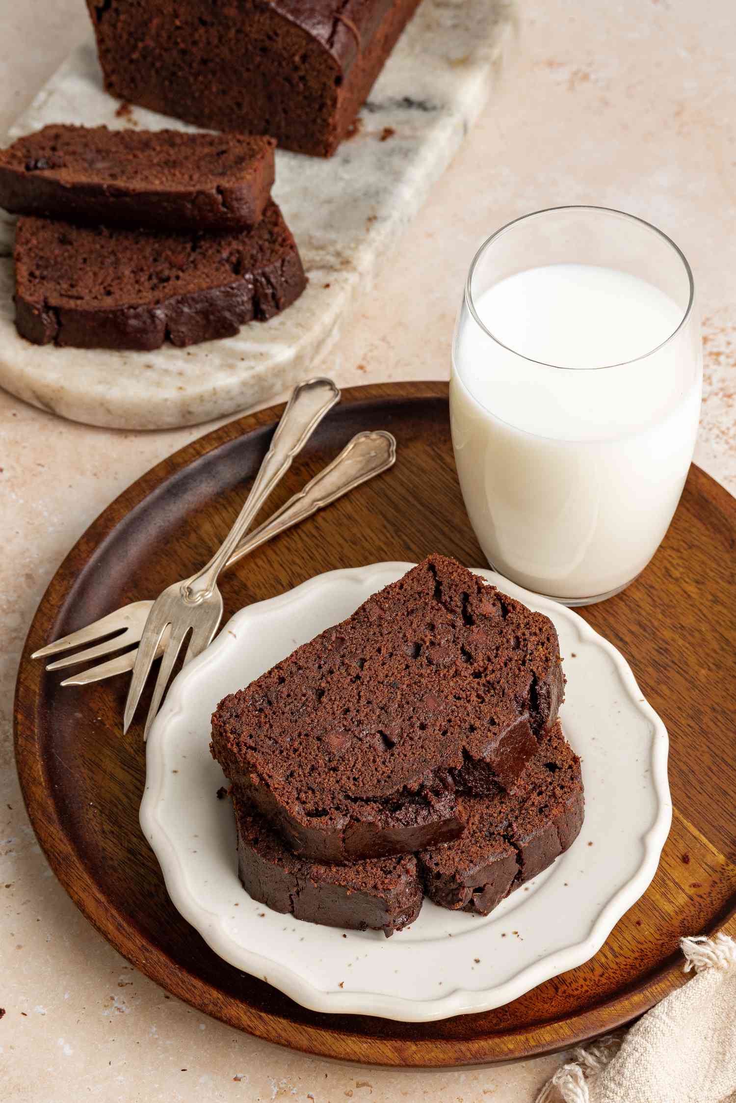 Chocolate Banana Bread Slices on a Small Plate Next to Two Forks and a Glass of Milk, All Sitting on a Circular Wooden Tray, and in the Background, a Marble Slab with More Slices of Bread