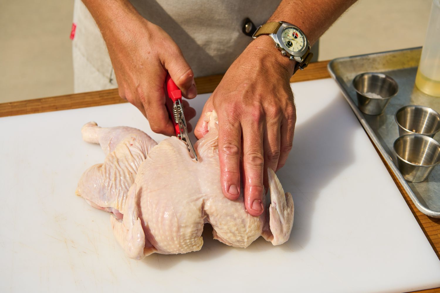 A person cutting a raw chicken in half with kitchen shears on a cutting board for Grilled Half Chicken recipe