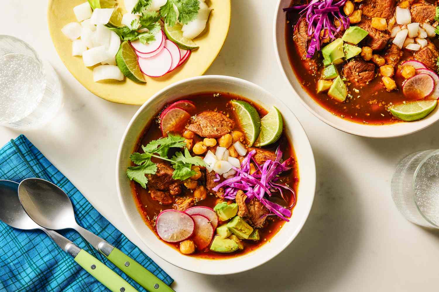 Two bowls of pozole rojo garnished with radishes lime avocado and purple cabbage