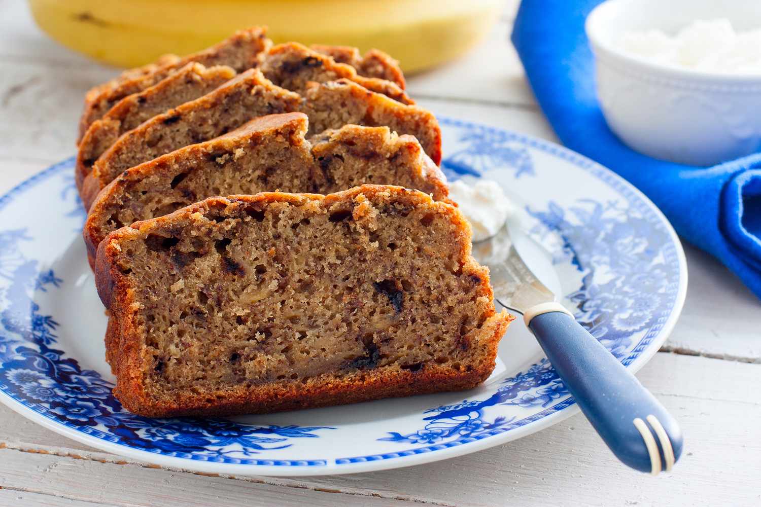 Sliced banana bread served on a decorative plate with a background of bananas and a blue napkin