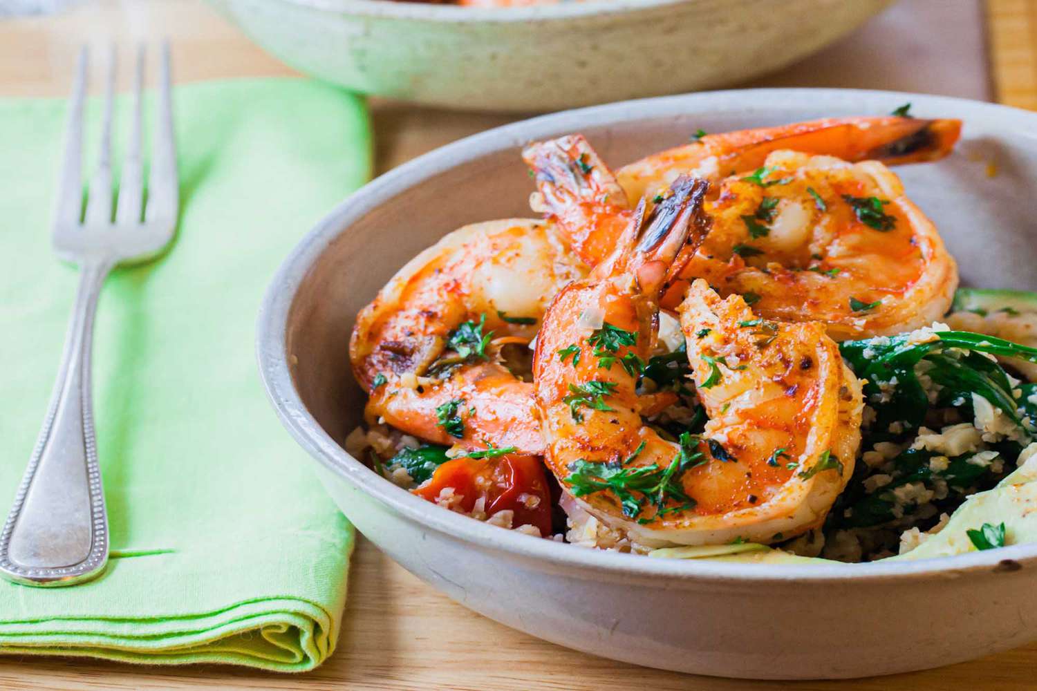 A shallow bowl of garlic shrimp cauliflower rice with a napkin and fork to the left. Sauteed shrimp and herbs are visible in the bowl.