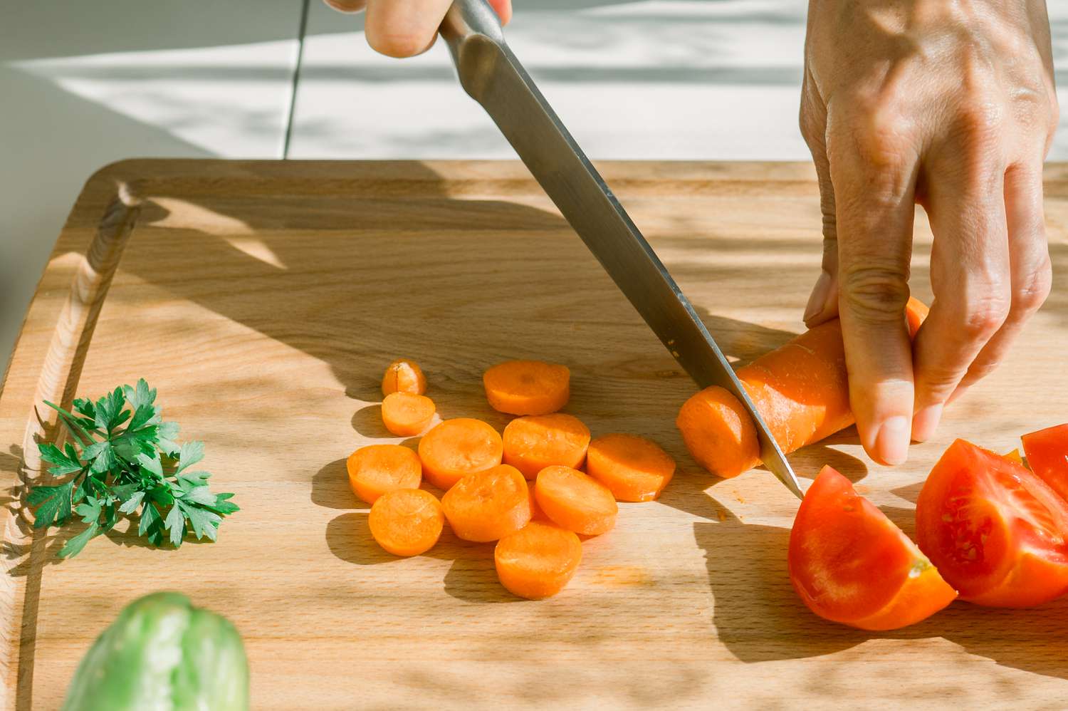 High angle of woman cutting carrot on wooden chopping board near tomatoes and parsley in kitchen during sunny weekend