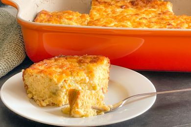 Angled view of a slice of Chile Cheese Egg Casserole on a white plate with a fork and the orange casserole dish in the background