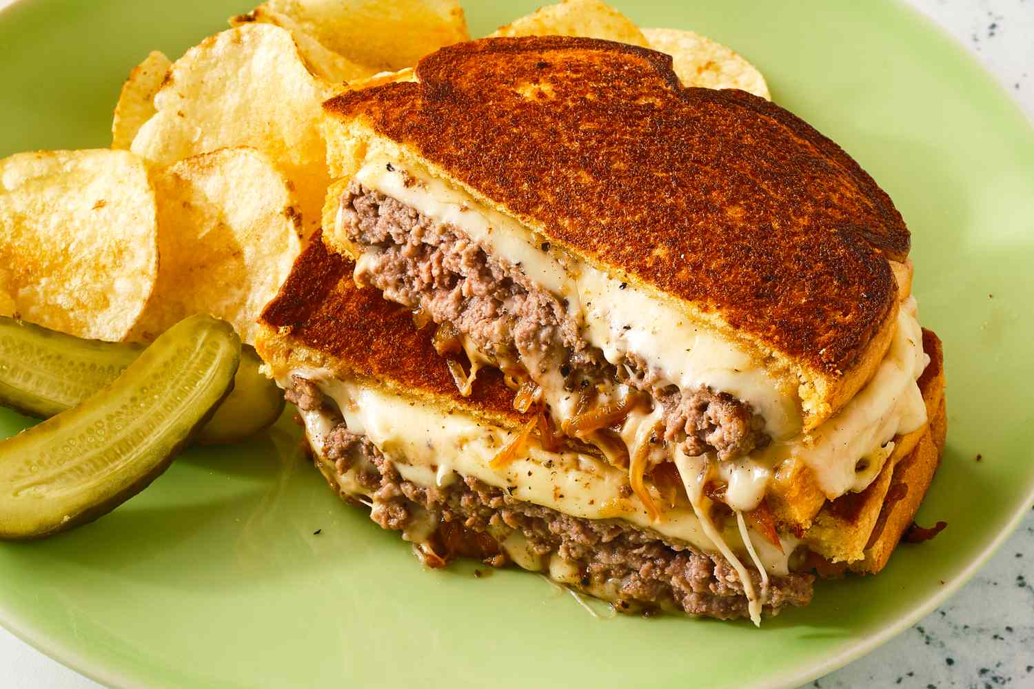 A patty melt with melted cheese and ground beef served with potato chips and pickles on a green plate