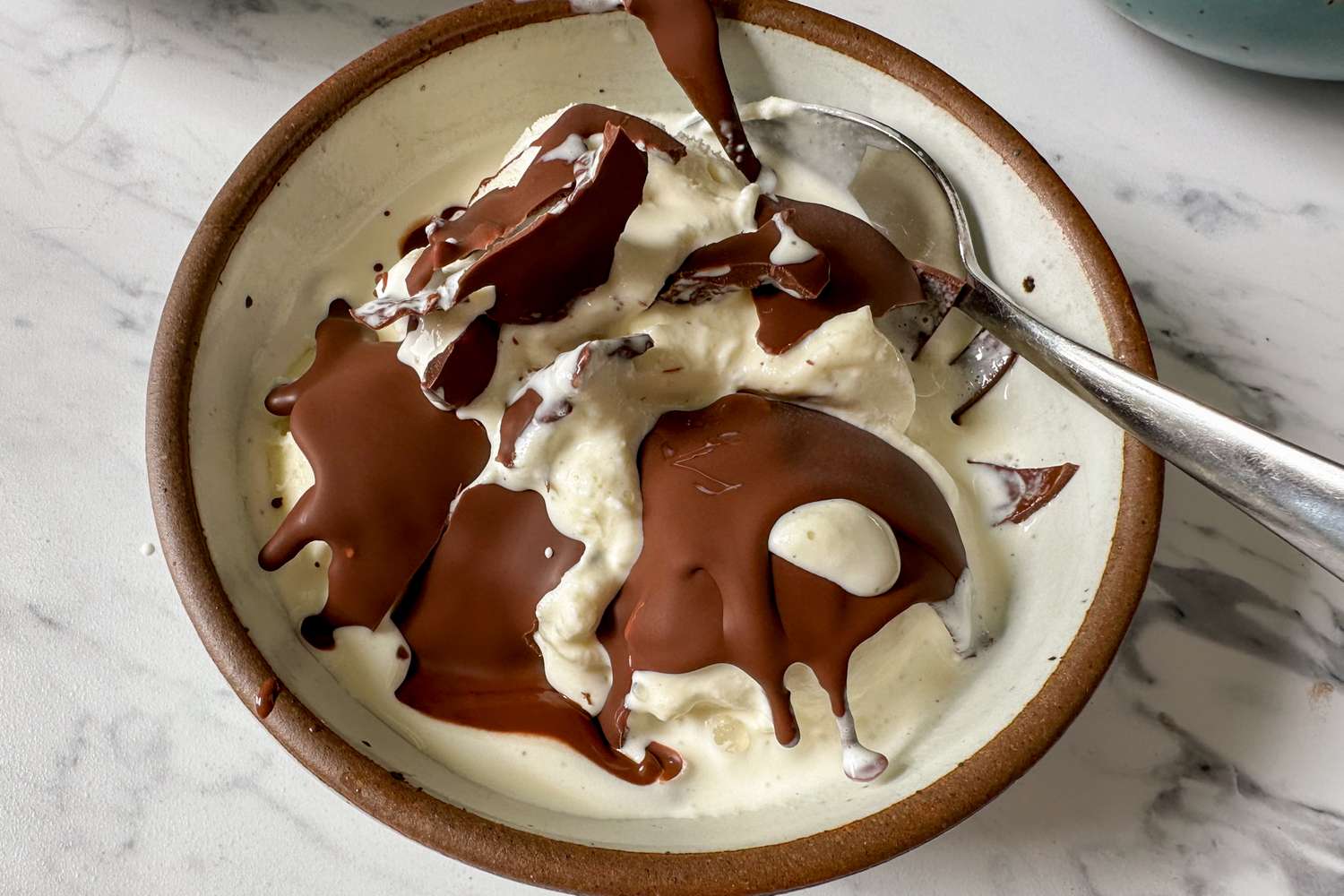 A bowl of ice cream topped with hardened chocolate shell with a spoon included in the bowl