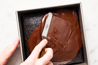 Overhead view of a hand using a spreader to frost over a single layer of chocolate cake in a square metal baking pan all on a marble countertop