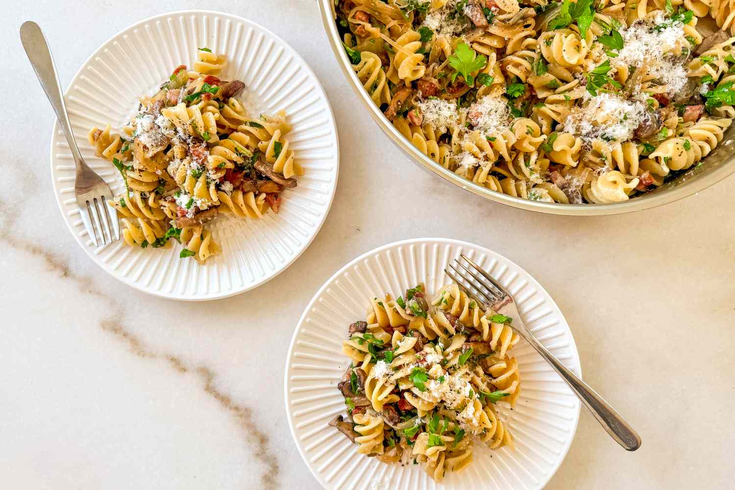 Plated servings of pasta with bacon and mushrooms garnished with parsley and Parmesan cheese served on white plates next to the cooking pan