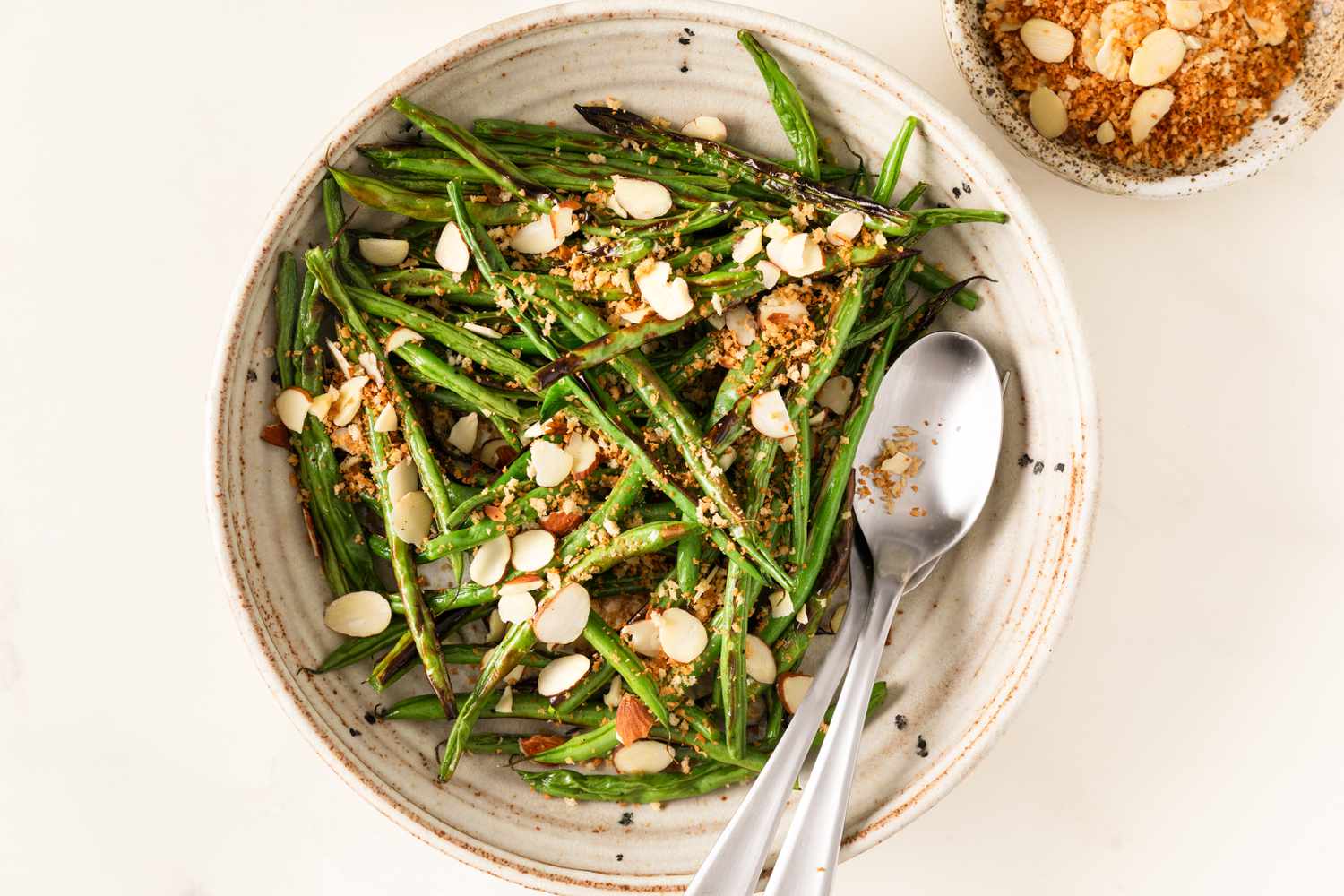 Bowl of broiled green beans topped with almond slices and crumbs, two spoons placed inside the bowl