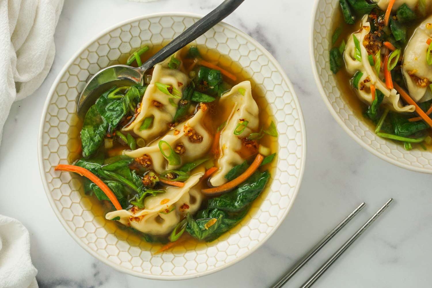 A bowl of dumpling soup garnished with vegetables and greens placed on a white surface with chopsticks beside