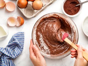 someone mixing chocolate cake batter in a bowl at a table with a carton of eggs, a glass of milk, a bowl of cocoa powder, and a bowl of flour