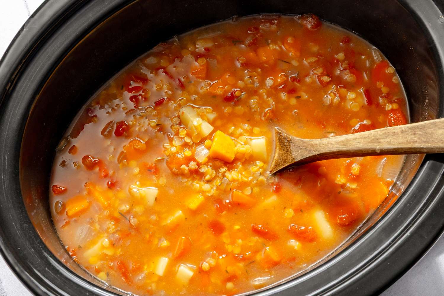 A slow cooker containing red lentil soup with vegetables and a wooden spoon