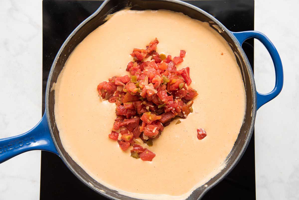 A black cast iron skillet will spices, milk and cheese being whisked together with a wooden spoon for easy homemade queso with tomatoes and rotel added in.