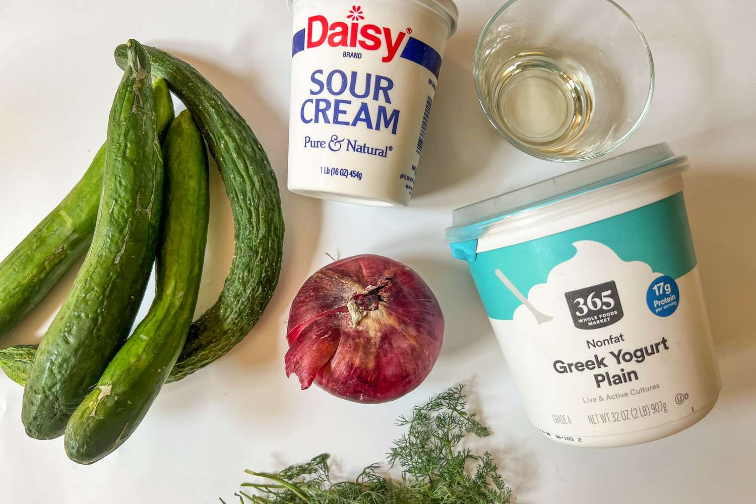 Ingredients for a cucumber salad on a surface, including cucumbers, a red onion, sour cream, Greek yogurt, and fresh dill
